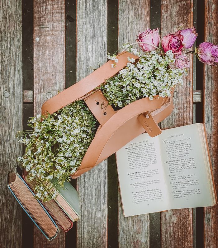hero-about Top view of an open book surrounded by a handbag filled with flowers on a wooden bench.
